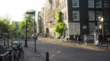 Amsterdam 'daki bulanık şehir manzarasında güneşli bir caddede yürüyen insanlar, Hollanda' da bisikletleri ve tarihi mimariyi yakalıyorlar..