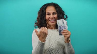 Sırıtan orta yaşlı bir kadın elinde kuyruğuyla mavi arka planda 1000 banknot tutuyor, zenginliği ve davetiyeyi simgeliyor..