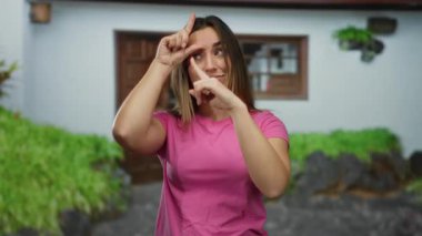 Genç bir kadın canlı bir sokak ortamında beceriksiz el hareketi yapıyor, pembe bir tişört giyiyor bulanık bir binanın arka planında..