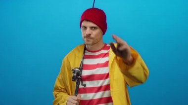 Sarı ceketli genç balıkçı mavi arka planda oltayla selam veriyor. Yakışıklı, kendine güvenen bir gülümsemesi var.