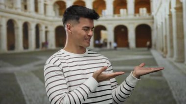 Çizgili süveter giymiş genç İspanyol bir adam tarihi bir üniversite avlusunda elini göstererek odak noktasını ve nişanı gösteriyor..