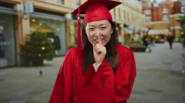 Kırmızı mezuniyet elbisesi giymiş kadın şehir sokağında sessiz hareketler yapıyor. Şenlikli dekorasyonlar ve gündüz vakti arka planda insanlar var.