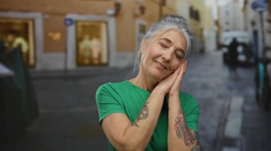 Gri saçlı, gülümseyen ve ellerini sokakta gezdiren, açık havada dövmelerini gösteren son sınıf öğrencisi bir kadın..