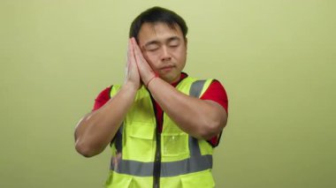 Güvenlik yeleği giymiş genç adam sarı arka planda uyuyor gibi görünüyor. Bu da yorgunluk ya da çalışma ortamında dinlenme hissi veriyor..