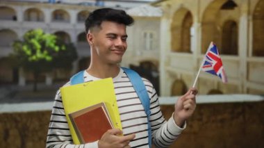 Eski bir üniversite avlusunda İngiliz bayrağı ve klasörleri taşıyan genç adam, eğitimi ve uluslararası çalışmaları sembolize ediyor.