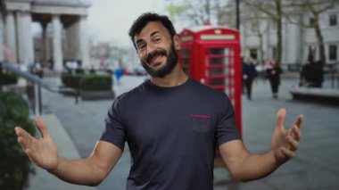 Sakallı İspanyol bir adam klasik İngiliz telefon kulübesi ve arka planda bulanık yayalar olan bir caddenin önünde durup ikonik bir şehir manzarası yakalıyor..