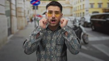 Şaşkın bakışlı İspanyol genç adam renkli binalar ve park edilmiş arabalarla şehir merkezinde şık gözlükler takıyor..