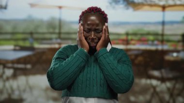 Terasta üzgün bir kadın, elleri yüzün üzerinde yeşil kazak giyiyor. Restoranın arka planında güneşli bir günde bulanık sandalyeler ve ağaçlar var.