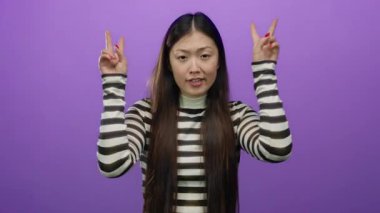 Tırnak işareti yapan bir kadın mor bir arka plana karşı iki eliyle hareket ediyor, çizgili gömlek giyiyor, alaycılık ya da ironi yapıyor, uzun saçlı ve gündelik ifadeli..