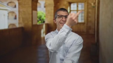 Eski bir üniversitede beyaz bir adam yaramazca gülümserken uygunsuz bir el hareketi yapıyor ve güneşli koşullar altında alışılmadık ifadeleri vurguluyor..