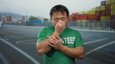 Yeşil gönüllü gömlekli genç adam arka planda nakliye konteynırlarıyla deniz kenarındaki limanda burnunu kapatıyor. Bu da deniz kenarında güçlü bir koku sorunu olduğunu gösteriyor..