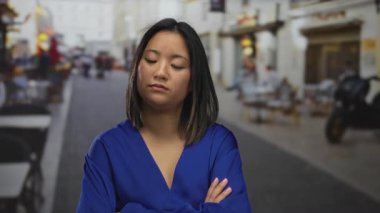 Şehir caddesinde kolları çapraz duran, mavi bir bluz giyen, şehir dışında gün ışığında düşünceli bir yüz ifadesi gösteren bir kadın..