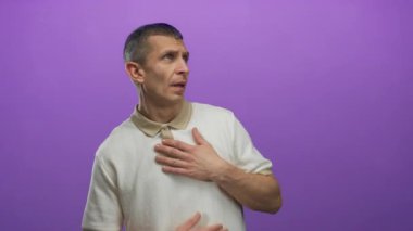 Beyaz gömlekli beyaz adam göğsünü mor arka planda tutuyor, stüdyo ortamında duygularını dışa vuruyor, merak ya da şaşkınlık hissi veriyor..