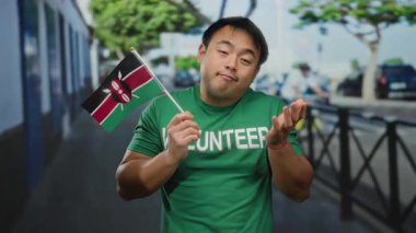 Kenya bayrağı taşıyan yeşil gönüllü gömlekli genç adam arka planda ağaçlar ve binalar olan şehir sokağında duruyor. Dışarıdaki uluslararası işbirliğini resmediyor..