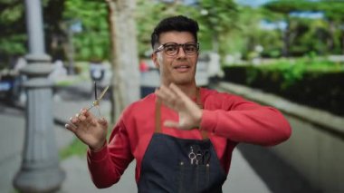 Genç İspanyol asıllı bir adam elinde makasla, kırmızı bir kazak ve önlükle dışarıda poz veriyor. Şehir arka planı hareketli ve davetkar bir atmosferi arttırıyor..