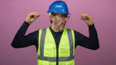 Şapkalı ve güvenlik yeleği olan İspanyol bir adam pembe arka planda komik bir yüz ifadesi takınıyor..