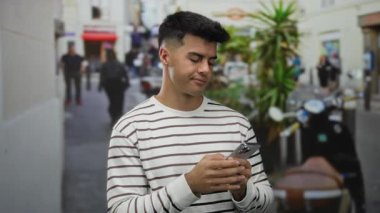 Şehir caddesinde insanlar ve bitkilerle çevrili akıllı telefon kullanan çizgili tişörtlü genç adam, şehir yaşam tarzını ve teknolojiyi modern cihazlarla açık havada gösteriyor.