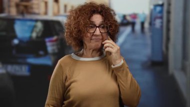 Gözlüklü İspanyol kadın şehir caddesinde akıllı telefon kullanıyor. Açık hava ortamında incelikle el kol hareketi yapıyor..