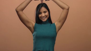 Genç bir kadın başının üstünde bir kalp oluşturur, sıcak kahverengi bir duvara gülümser, basit ve izole bir ortamda sevgi ve mutluluk hissini resmeder..