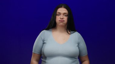 Uzun saçlı genç bir kadın, açık mavi bir gömlek giyerken hayal kırıklığını ya da stresi ifade eden mavi bir arka planda parmaklarıyla bir jest yapar..