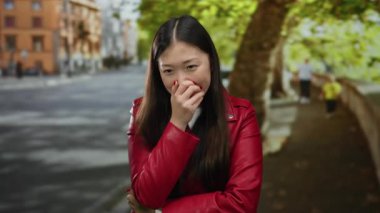 Kırmızı ceketli kadın, arka planda ağaçlar ve binalarla şehir caddesinde gülümsüyor.