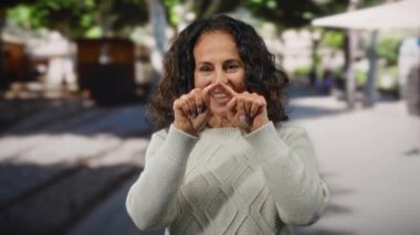 Kıvırcık saçlı bir kadın şehir ortamında gülümserken beyaz bir kazak giyer ve hareketli bir sokak ortamında pozitif bir hava izlenimi verir..