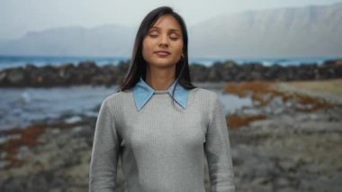 Gri kazaklı kadın, gözleri kayalık deniz kenarında kapalı duruyor. Yumuşak, bulutlu bir gökyüzünün altında okyanusun dinginliğini kucaklıyor..