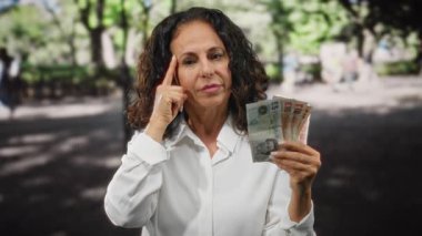 Orta yaşlı bir kadın, İngiliz banknotlarını açık havada tutuyor. Düşünüyor ve bir şehir caddesini işaret ediyor..