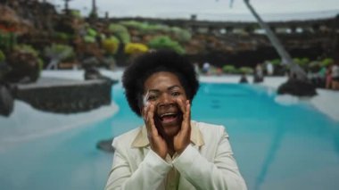 Beyaz ceketli bir kadın, canlı yeşilliklerle çevrili lüks bir tatil köyüne neşe içinde bağırıyor, dışarıda bir an heyecan ve mutluluk yakalıyor..