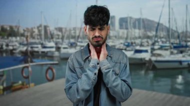 Boğazı ağrıyan genç bir adam arka planda tekneler ve su olan bir limanın yanında duruyor..