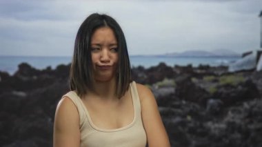 Sahilde arka planda okyanus ve bulutlu gökyüzü olan genç bir kadın, kollarını kavuşturmuş düşünceli ve düşünceli görünüyor, temiz havanın ve doğal manzaranın tadını çıkarıyor..