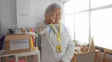 Gönüllü üniformalı gri saçlı bir kadın davetkar bir hayır kurumuna davetkar bir şekilde el kol hareketi yapıyor. Kıyafetleri ve kutuları görünür..