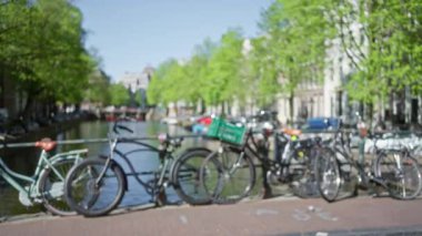 Bulanık bisikletler, berrak mavi gökyüzü altında çözünmüş yeşillik ve tarihi mimari ile Amsterdam Kanalı boyunca dizilmiş sakin bir şehir manzarası yaratıyor..