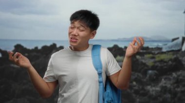 Sırt çantalı genç adam, kayalık sahil kenarında el kol hareketi yapıyor. Sahilde rahat bir öğrenci havası var. Bulutlu bir günde açık havada, deniz kenarında bir anı yakalıyor..