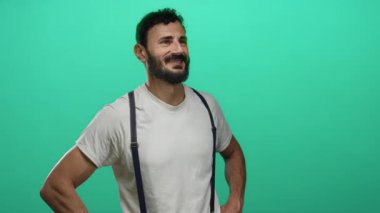 Sakallı İspanyol sakallı, elleri kalçasında canlı yeşil bir arka plana yaslanmış bir şekilde gülümseyen, stüdyo ortamında günlük beyaz gömlek ve pantolon askısı takan bir adam.