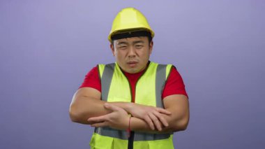 Kask ve güvenlik yeleği giyen genç adam, izole edilmiş mor bir arka plan önünde soğuk görünüyor. Rahatsızlık ve ürperti hislerini iletiyor..