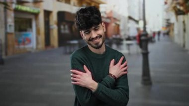 Bir kenar mahallede kendini kucaklayan genç adam, gülümseyerek kendini sevgiyle ve sükunetle ifade ediyor. Arka planda iki adam ve parlak ışık, kalabalık bir şehirde huzurlu bir anı yakalıyor.