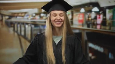 Mezuniyet giysisi içindeki genç bir kadın kalabalık bir alışveriş merkezinde neşeli bir şekilde gülümsüyor, mağaza tabelalarının arasında başarı ve mutluluğu somutlaştırıyor ve evin içinde hareketli bir atmosfer yaratıyor..
