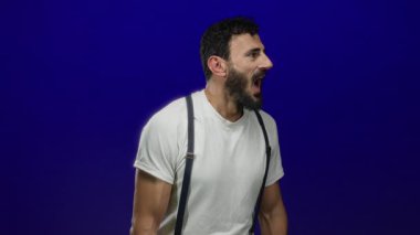 Beyaz tişörtlü ve pantolon askısı takmış İspanyol bir adam dramatik bir etki yaratmak için mavi arka planda sürpriz yapıyor.