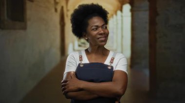 Önlük takmış, kollarını kavuşturmuş kendinden emin bir kadın kemerli koridorları olan eski bir üniversitede kapalı bir yerde duruyor ve gururunu ve güvenini dile getiriyor..