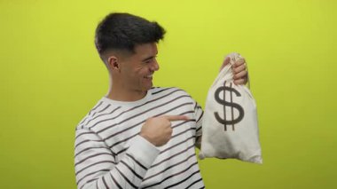 Genç İspanyol bir adam gülümsüyor ve para çantasına canlı sarı bir arkaplan üzerinden işaret ediyor, neşeli bir ifade ve finansal başarı sergiliyor..