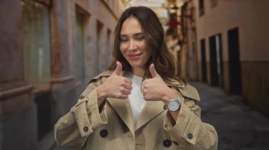 Bir şehir caddesinde başparmak kaldıran genç bir kadın, pozitifliğini ve özgüvenini dışa vuruyor, mutlu ve neşeli bir tavırla şehir ortamında bej renkli bir trençkot giyiyor..