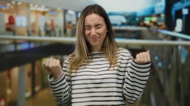 Çizgili tişörtlü genç bir kadın kapalı bir alışveriş merkezinde dikilirken güvenle gülümsüyor..