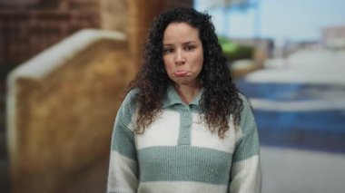 Sokakta çizgili kazak giyen kıvırcık saçlı İspanyol kadın şehir ortamında üzüntü ifade ediyor..