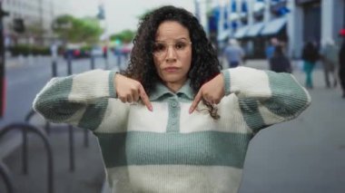Yoğun caddede aşağı doğru hareket eden bir kadın şehir ortamında ciddi bir ifadeyle yön veya konumu belirterek kentsel ve gündelik tarzı harmanlıyor.