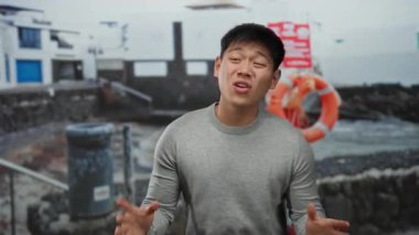 Gri kazaklı genç adam sahilde duygularını ifade ediyor. Arka planda can yelekleri ve binalar var. Sahilde bir açık hava sahnesi sergiliyor..