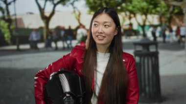 Kırmızı ceketli uzun saçlı kadın, güneşli bir günde ağaçlarla ve arka planda insanlarla şehir sokağında dikilirken miğfer tutuyor..