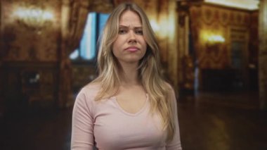 Genç sarışın kadın otelin lobisinde kamerayı göstererek sert bir ifade ve dik dik bakışıyla gerilimi kınıyor..