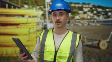 Yansıtıcı yelek giymiş genç İspanyol bir adam deniz kenarında bir plajda tablet tutuyor. İnşaat uzmanlığını doğanın güzelliğiyle harmanlıyor..