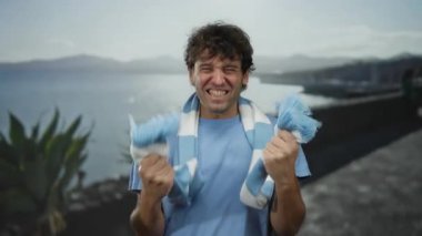 İspanyol adam güneşli bir sahilde futbol zaferini kutluyor çarpıcı bir deniz zeminine karşı çizgili bir eşarp sergiliyor tutku, heyecan ve spor ruhunu yansıtıyor..